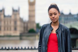 A young black woman in a red camisole and black leather jacket with her hait up in a bun stands in front of a blurred house of parliament 