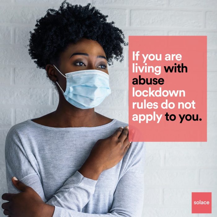 A picture of a black girl with curly hair up in a puff with a standard surgical mmask on looking closed off. She is wearing grey standing against a grey wall
