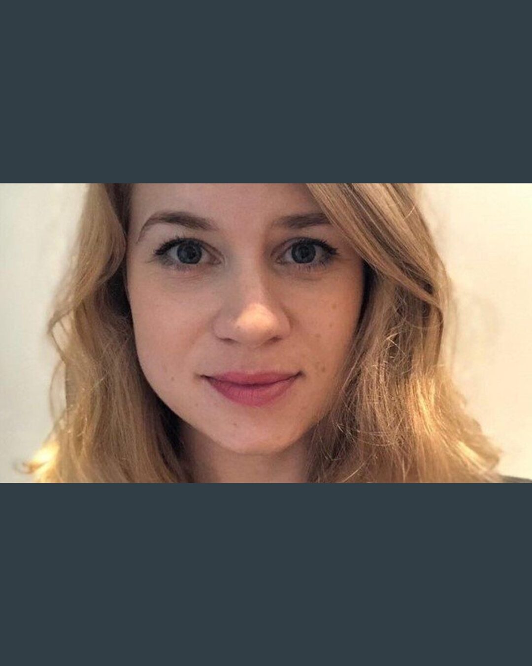Close-up portrait of a young woman with shoulder-length blonde hair, fair skin, and blue eyes, looking directly at the camera with a neutral expression against a plain indoor background.