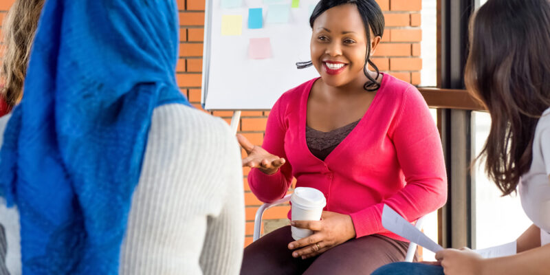 A woman in a pink jumper leads a small group discussion, smiling and gesturing while holding a coffee cup, as several participants sit around her in a meeting space with a whiteboard covered in colourful sticky notes behind her.