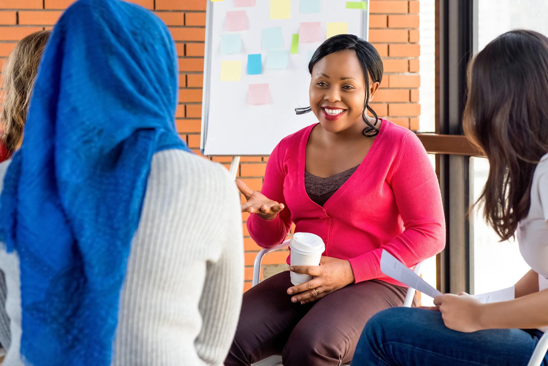 A woman in a pink jumper leads a small group discussion, smiling and gesturing while holding a coffee cup, as several participants sit around her in a meeting space with a whiteboard covered in colourful sticky notes behind her.